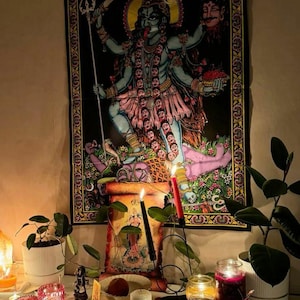 May include: A black tapestry depicting the Hindu goddess Kali, with multiple arms and a fierce expression. The image is framed by a decorative border. The scene includes candles, plants, and other spiritual items on a white surface, creating a devotional atmosphere.