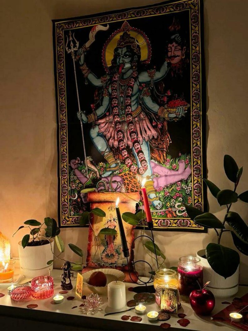 May include: A black tapestry depicting the Hindu goddess Kali, with multiple arms and a fierce expression. The image is framed by a decorative border. The scene includes candles, plants, and other spiritual items on a white surface, creating a devotional atmosphere.