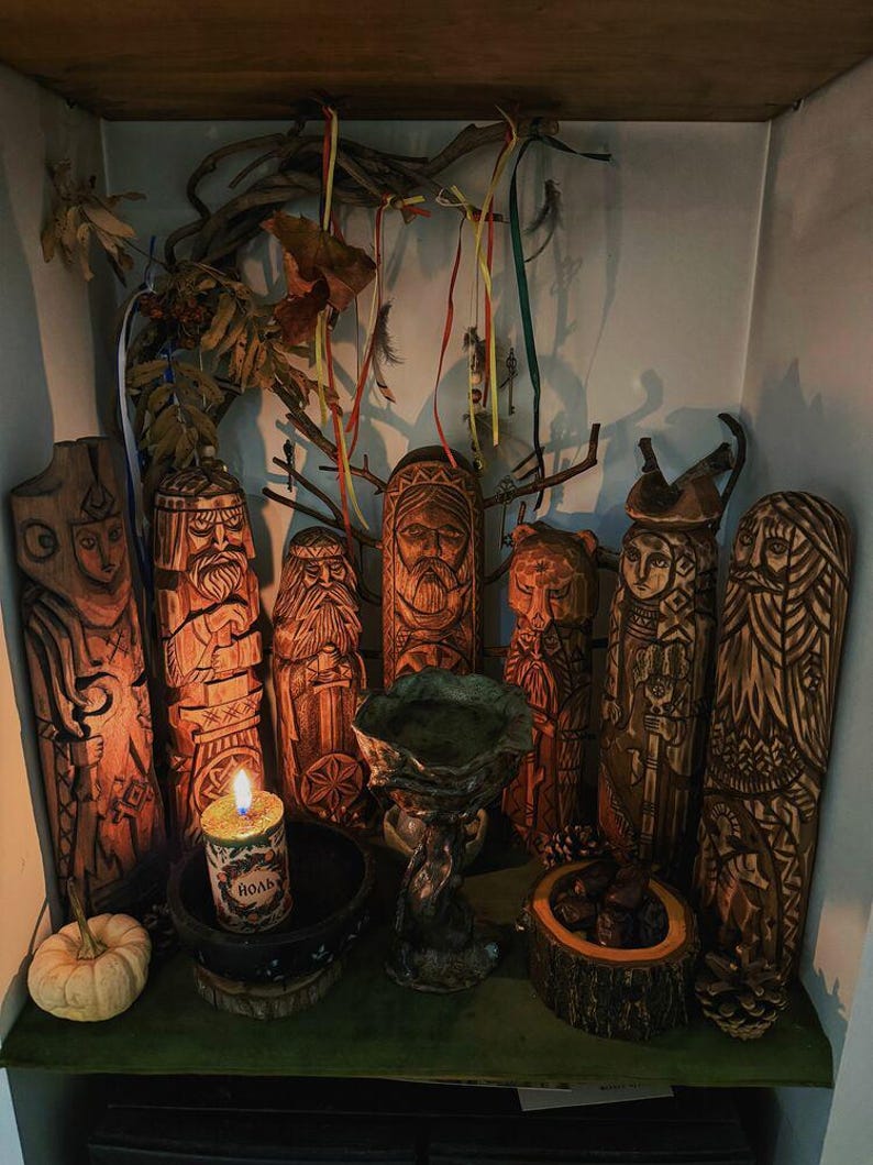May include: A collection of hand-carved wooden figures with intricate details, arranged on a shelf. The figures depict various faces and forms, with a central candle and small pumpkin. Decorative branches and ribbons add to the display.