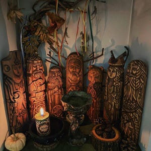 Puede incluir: Una colecci&oacute;n de figuras de madera talladas a mano con detalles intrincados, dispuestas en una estanter&iacute;a. Las figuras representan varios rostros y formas, con una vela central y una peque&ntilde;a calabaza. Ramas decorativas y cintas a&ntilde;aden a la exhibici&oacute;n.