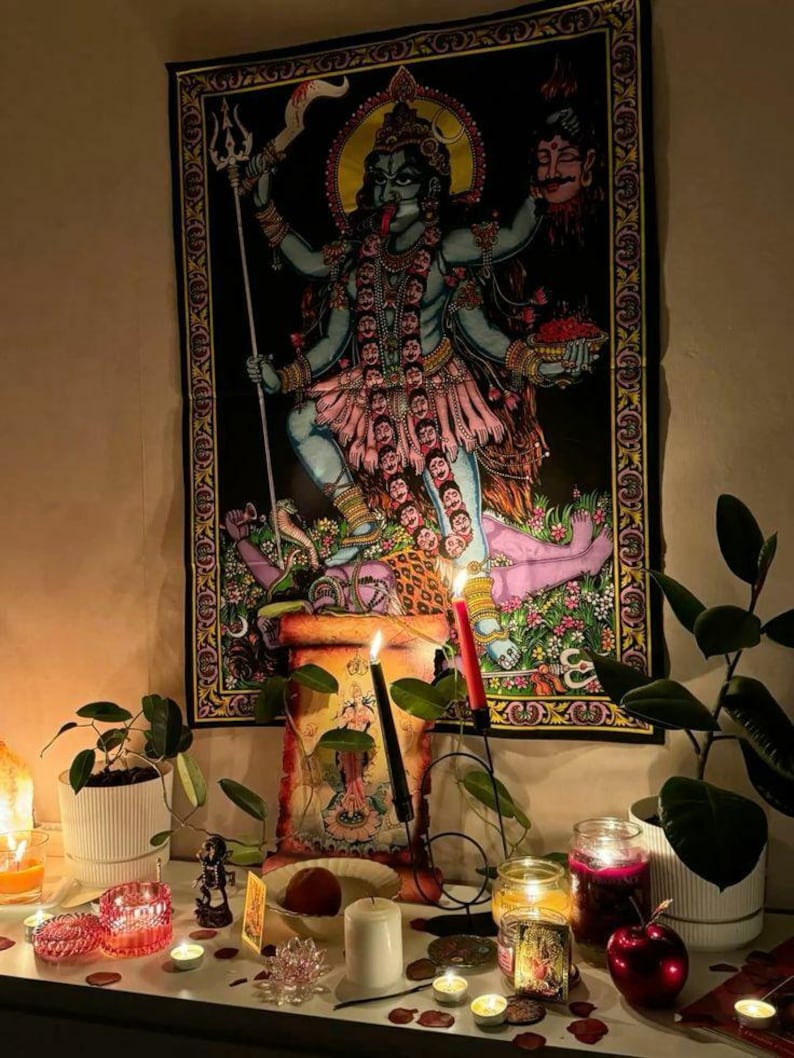 Puede incluir: Un tapiz negro que representa a la diosa hind&uacute; Kali, con m&uacute;ltiples brazos y una expresi&oacute;n feroz. La imagen est&aacute; enmarcada por un borde decorativo. La escena incluye velas, plantas y otros objetos espirituales sobre una superficie blanca, creando una atm&oacute;sfera devocional.