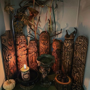 May include: A collection of hand-carved wooden figures with intricate details, arranged on a shelf. The figures depict various faces and forms, with a candle, small pumpkin, and decorative branches adding to the display. The figures are in shades of brown.