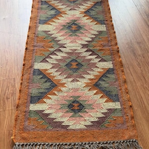 May include: A rectangular woven runner rug featuring a repeating geometric diamond pattern in shades of orange, cream, sage green, and dark gray. The rug has a textured surface and is finished with a fringe of loose threads along the bottom edge.