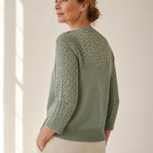 May include: A pale green knitted cardigan featuring an openwork pattern on the upper back and sleeves. The cardigan has three-quarter length sleeves and a ribbed hem. The model is wearing beige trousers.