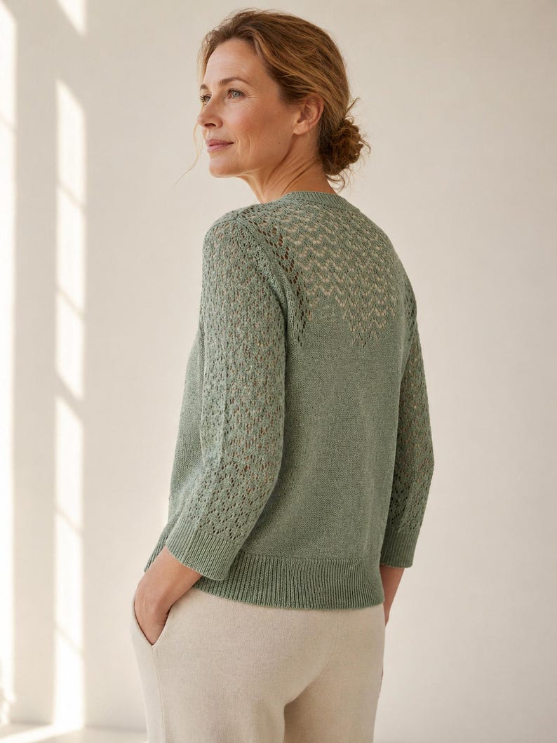 May include: A pale green knitted cardigan featuring an openwork pattern on the upper back and sleeves. The cardigan has three-quarter length sleeves and a ribbed hem. The model is wearing beige trousers.