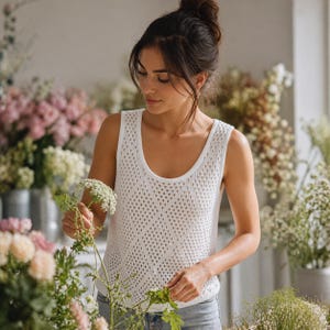 Puede incluir: Un top sin mangas blanco de punto abierto con cuello redondo. La parte superior la lleva una persona en una florister&iacute;a, rodeada de varias flores en recipientes met&aacute;licos. La persona sostiene una ramita de flores blancas.