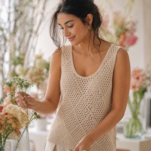 May include: A beige, open-knit tank top with a scoop neck. The top is worn by a person arranging flowers in a vase. The background features a variety of colorful flowers and soft lighting, creating a natural and serene atmosphere.