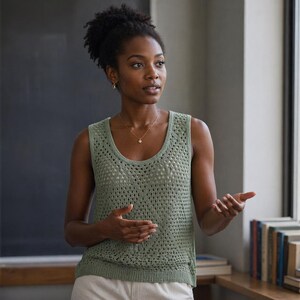 May include: A woman wearing a light green, open-knit tank top. She is wearing a gold necklace and earrings. The background includes a chalkboard, a window, and a wooden shelf with books.