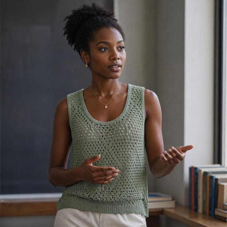 May include: A woman wearing a light green, open-knit tank top. She is wearing a gold necklace and earrings. The background includes a chalkboard, a window, and a wooden shelf with books.