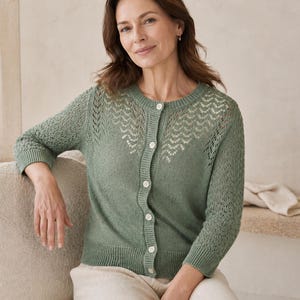 May include: A sage green cardigan with a detailed openwork pattern across the upper chest and sleeves. The button-down sweater has three-quarter sleeves and is paired with cream-coloured trousers. The cardigan is made of a soft knit material.