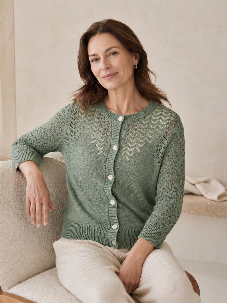 May include: A sage green cardigan with a detailed openwork pattern across the upper chest and sleeves. The button-down sweater has three-quarter sleeves and is paired with cream-coloured trousers. The cardigan is made of a soft knit material.