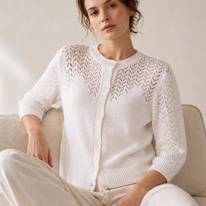 May include: A white, open-knit cardigan with a round neckline and three-quarter sleeves. The cardigan features a delicate, openwork design across the upper chest and sleeves. The model is wearing matching white pants.
