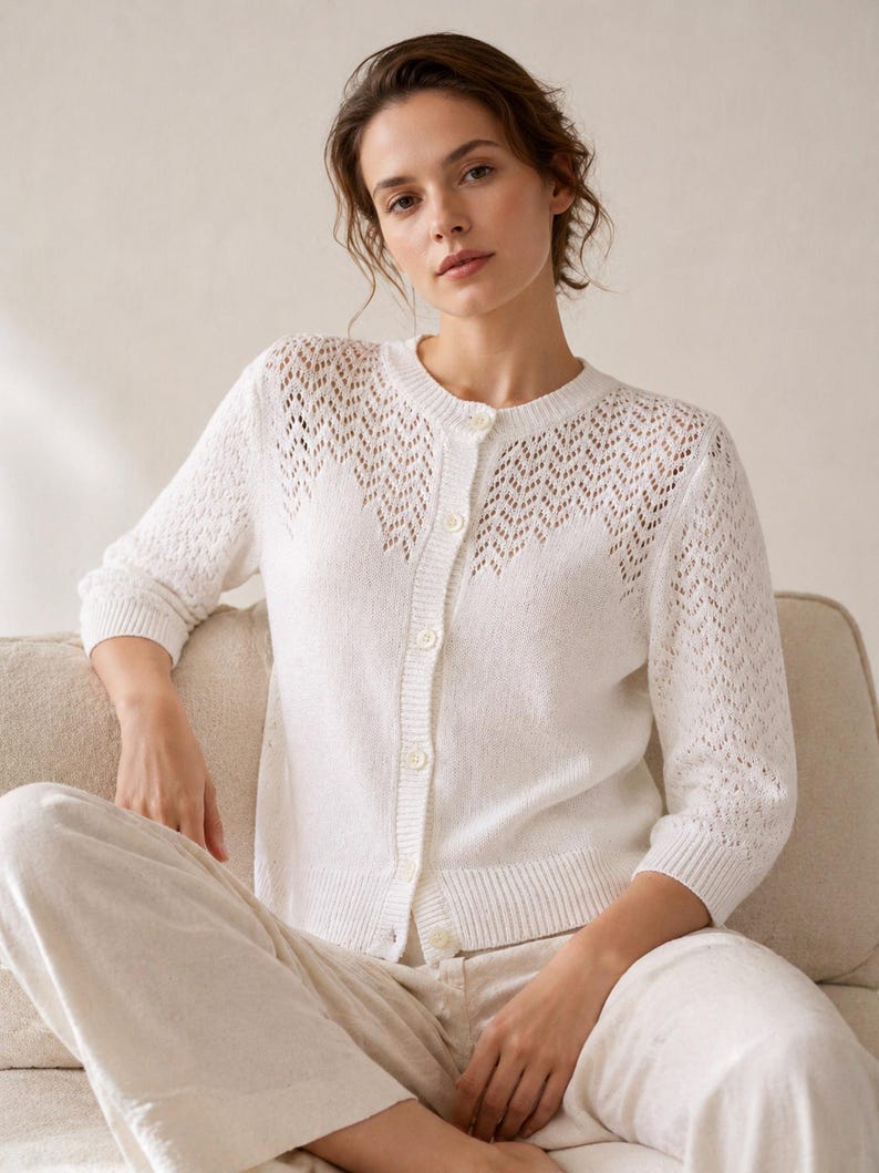 May include: A white, open-knit cardigan with a round neckline and three-quarter sleeves. The cardigan features a delicate, openwork design across the upper chest and sleeves. The model is wearing matching white pants.