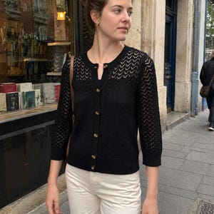 Op de afbeelding: Een zwarte cardigan met een knoopsluiting en een delicaat open breipatroon op de bovenborst en mouwen. De cardigan wordt gecombineerd met een cr&egrave;mekleurige broek. De outfit wordt buiten gedragen.