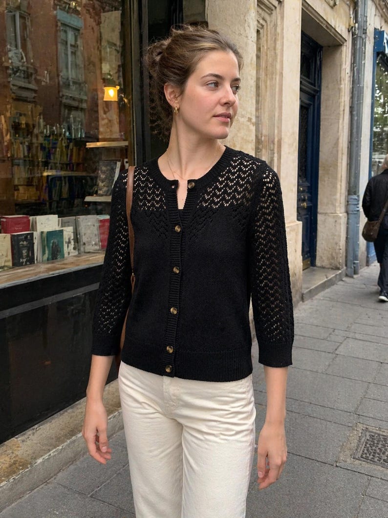 Op de afbeelding: Een zwarte cardigan met een knoopsluiting en een delicaat open breipatroon op de bovenborst en mouwen. De cardigan wordt gecombineerd met een cr&egrave;mekleurige broek. De outfit wordt buiten gedragen.