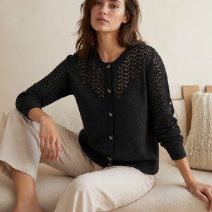 Op de afbeelding: Een zwarte cardigan met knoopsluiting en een delicaat openwerkontwerp over de bovenborst en mouwen. De cardigan wordt gecombineerd met lichtgekleurde broeken. Het model zit op een neutraal getinte bank.