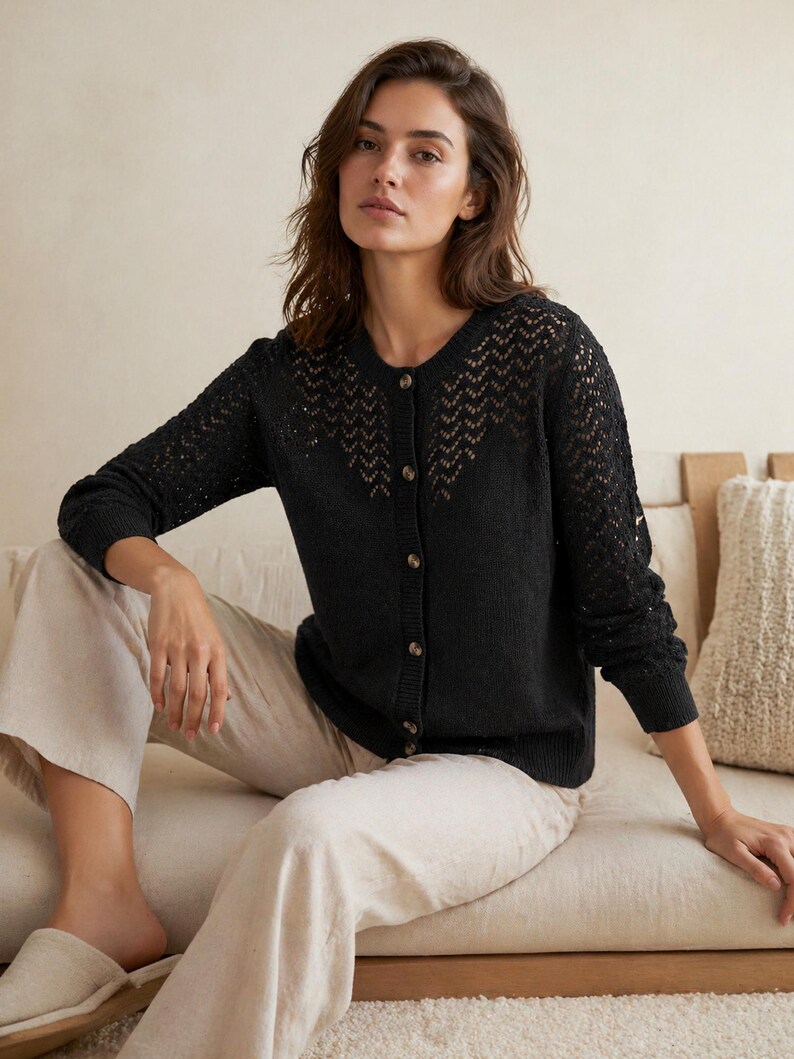 Op de afbeelding: Een zwarte cardigan met knoopsluiting en een delicaat openwerkontwerp over de bovenborst en mouwen. De cardigan wordt gecombineerd met lichtgekleurde broeken. Het model zit op een neutraal getinte bank.