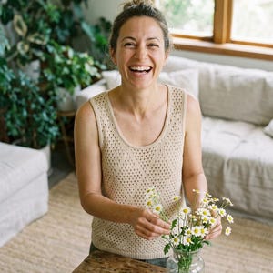 May include: A woman smiles as she arranges daisies in a clear glass vase on a wooden table. She wears a beige, open-knit tank top. The room has a neutral colour palette with a sofa and plants in the background.