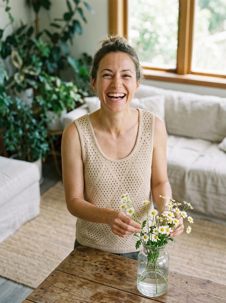 May include: A woman smiles as she arranges daisies in a clear glass vase on a wooden table. She wears a beige, open-knit tank top. The room has a neutral colour palette with a sofa and plants in the background.