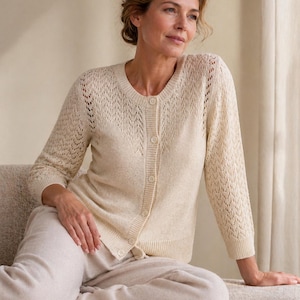 May include: A cream-coloured cardigan with a button-up front and openwork detailing on the sleeves. The cardigan is paired with matching light beige trousers. The outfit is made of a soft, knit material.