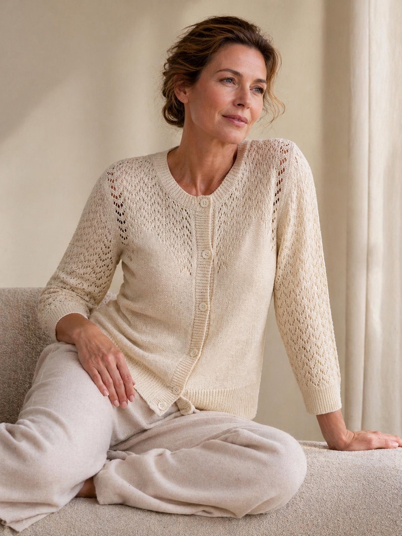 May include: A cream-coloured cardigan with a button-up front and openwork detailing on the sleeves. The cardigan is paired with matching light beige trousers. The outfit is made of a soft, knit material.