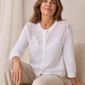 May include: A white, button-down cardigan with a delicate, open-knit design on the upper chest and sleeves. The cardigan has a round neckline and three-quarter sleeves. The model is wearing beige pants.