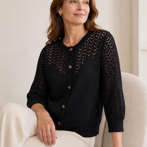 May include: A black knitted cardigan with a round neck and three-quarter sleeves. The garment has an openwork pattern on the upper chest and sleeves, and a button-down front. The cardigan is worn with cream-coloured trousers.