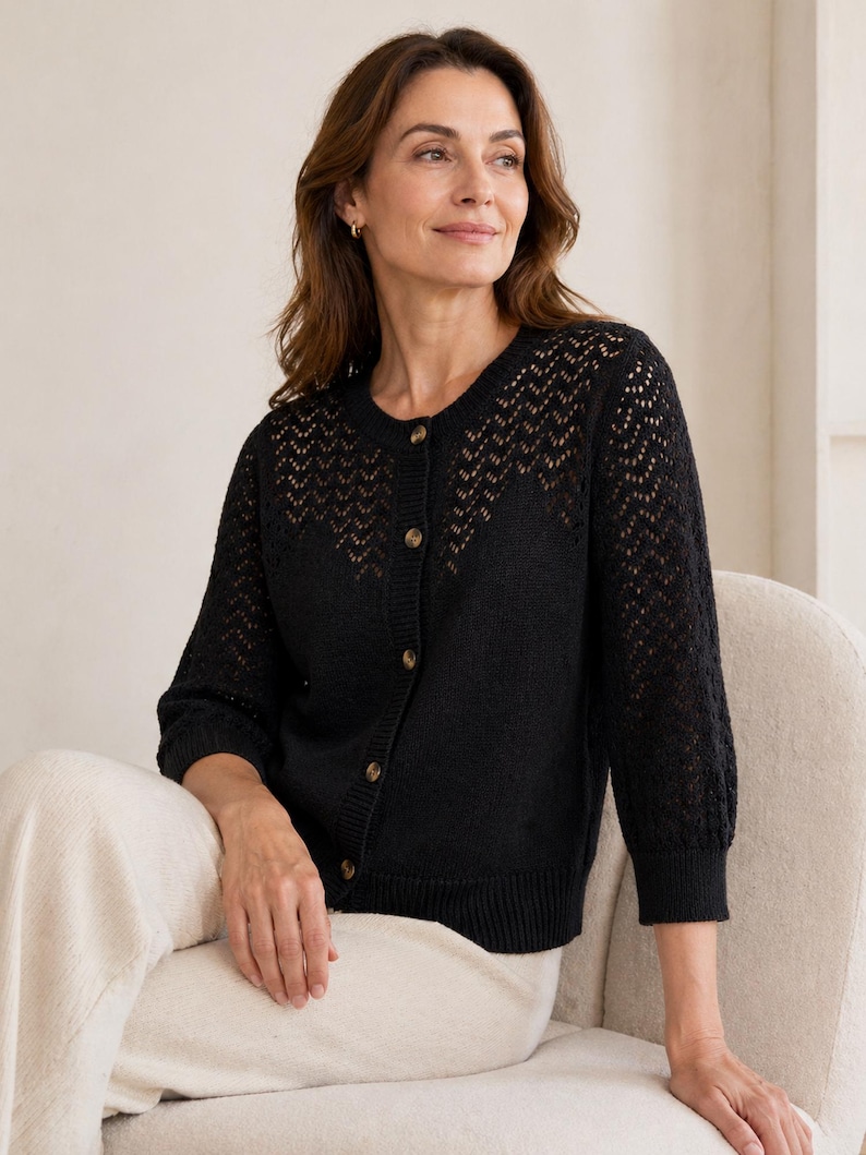 May include: A black knitted cardigan with a round neck and three-quarter sleeves. The garment has an openwork pattern on the upper chest and sleeves, and a button-down front. The cardigan is worn with cream-coloured trousers.