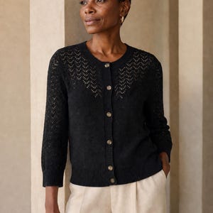 May include: A black cardigan with a round neckline and button-up front. The cardigan has an openwork design on the shoulders and sleeves. The model is wearing the cardigan with cream-coloured trousers.