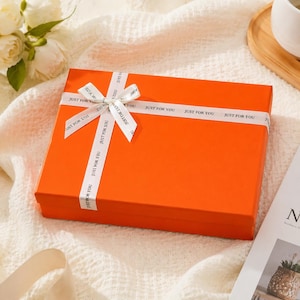 May include: A bright orange gift box, tied with a white ribbon printed with "JUST FOR YOU". The box is rectangular and rests on a white surface. The ribbon is tied in a bow.