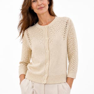 May include: A cream-coloured cardigan with a round neckline and button-down front. The sleeves are three-quarter length, and the shoulders feature a decorative open-knit pattern. The cardigan is paired with light-coloured trousers.