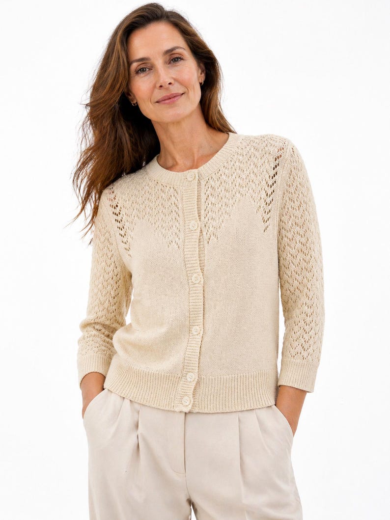May include: A cream-coloured cardigan with a round neckline and button-down front. The sleeves are three-quarter length, and the shoulders feature a decorative open-knit pattern. The cardigan is paired with light-coloured trousers.