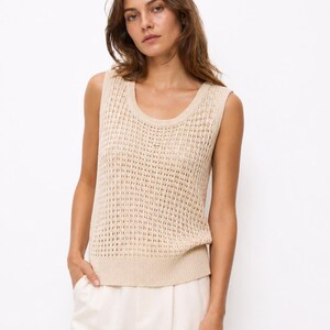 Puede incluir: Un top sin mangas de punto beige con cuello redondo. El top tiene un dise&ntilde;o de tejido abierto. La modelo lleva el top con pantalones blancos. El fondo es blanco liso.