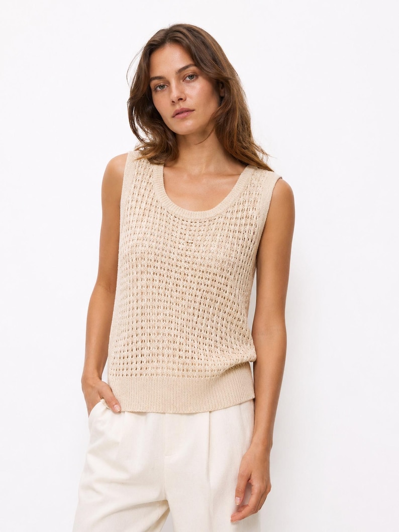 Puede incluir: Un top sin mangas de punto beige con cuello redondo. El top tiene un dise&ntilde;o de tejido abierto. La modelo lleva el top con pantalones blancos. El fondo es blanco liso.