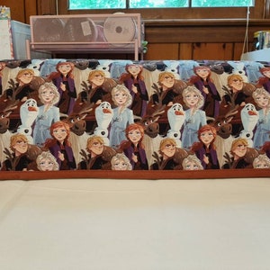 May include: A brown and orange fabric cover with a repeating pattern of characters from the movie Frozen. The characters include Elsa, Anna, Olaf, and Sven. The fabric is a soft, cotton blend.