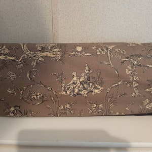 May include: A brown fabric with a black and white toile pattern featuring a scene of people and animals in a garden setting.