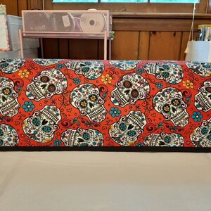 May include: Red fabric with a repeating pattern of white sugar skulls with floral designs. The skulls have black eyes and a black outline. The fabric has a black trim.