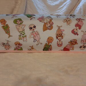 May include: White fabric with a pink border featuring a repeating pattern of bald women wearing various outfits and accessories. The women are depicted in a cartoon style with large eyes and bright colors.