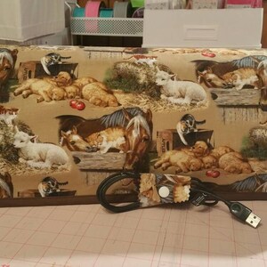 May include: A brown and tan fabric cover with a horse, cats, and puppies printed on it. The cover has a brown trim and a black cord with a brown fabric strap attached to it.