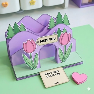 Puede incluir: Una tarjeta de felicitación desplegable con una escena de montaña lavanda, árboles verdes y tulipanes rosas. La tarjeta dice "MISS YOU" y "CAN'T WAIT TO SEE YOU". Un corazón rosa está al lado de la tarjeta. La tarjeta está sobre una superficie verde.