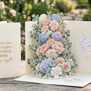 May include: A cream-coloured pop-up greeting card with a floral arrangement of pink, blue, and peach flowers. The card reads "To the one who makes everything bloom." A heart-shaped seed paper is in a pocket.