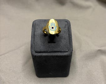 May include: A gold-toned ring featuring an oval-shaped face with a Hamsa hand symbol. The Hamsa design is outlined in light blue with a small, dark blue circular stone centred in the palm. The ring band is smooth and matches the gold-toned finish.
