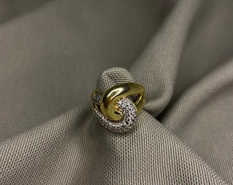 May include: A gold ring featuring an intertwined knot design. One half of the knot is a smooth, polished gold band, while the other half is encrusted with small, clear, sparkling stones that cover the surface in a dense, pavé-style arrangement.