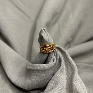 May include: A gold ring featuring a multi-band design with several small, round, vibrant red gemstones embedded throughout the metal structure. The ring has a textured, layered appearance with the red stones set at varying intervals along the bands.