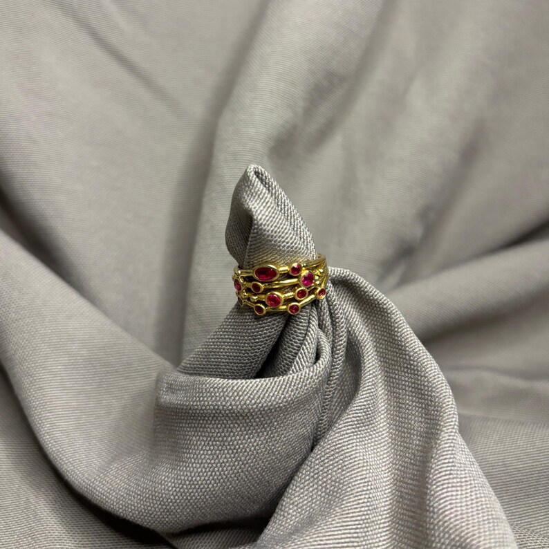 May include: A gold ring featuring a multi-band design with several small, round, vibrant red gemstones embedded throughout the metal structure. The ring has a textured, layered appearance with the red stones set at varying intervals along the bands.
