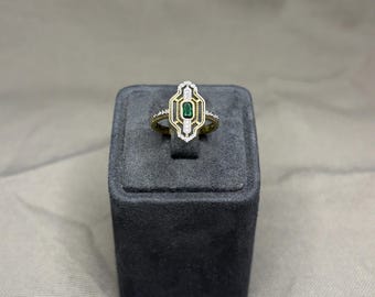 May include: A gold ring featuring an elongated, geometric face with a central rectangular green stone. The setting is accented with small, clear stones arranged in vertical lines, creating a symmetrical, multi-layered frame around the centre.