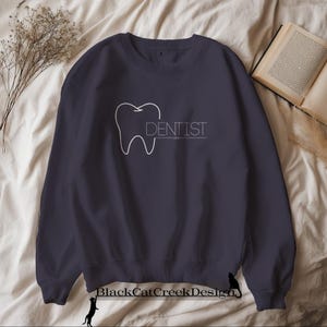 Peut inclure: Sweat-shirt bleu marine avec un dessin blanc d'une dent et le mot "DENTIST" imprimé en blanc. Le sweat-shirt est de style col rond et est fait d'une matière douce.
