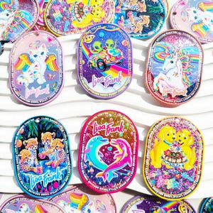 Pode incluir: Uma coleção de pingentes ovais com tema Lisa Frank. Cada pingente apresenta ilustrações coloridas de unicórnios, gatos, chitas, golfinhos e alienígenas. Os pingentes têm bordas brilhantes e o texto "Lisa Frank".