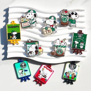 Peut inclure: Une collection de dix épinglettes en acrylique représentant Snoopy dans diverses scènes sur le thème du café, incluant la marque Starbucks, des panneaux en forme de chevalet avec le texte SNOOPY et des personnages avec des tasses.
