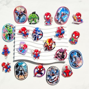 May include: A collection of colourful acrylic pins featuring various comic book superheroes including Spider-Man, Hulk, Batman, Iron Man, and Captain America in both chibi and detailed illustrated styles, arranged on a white wavy display stand.